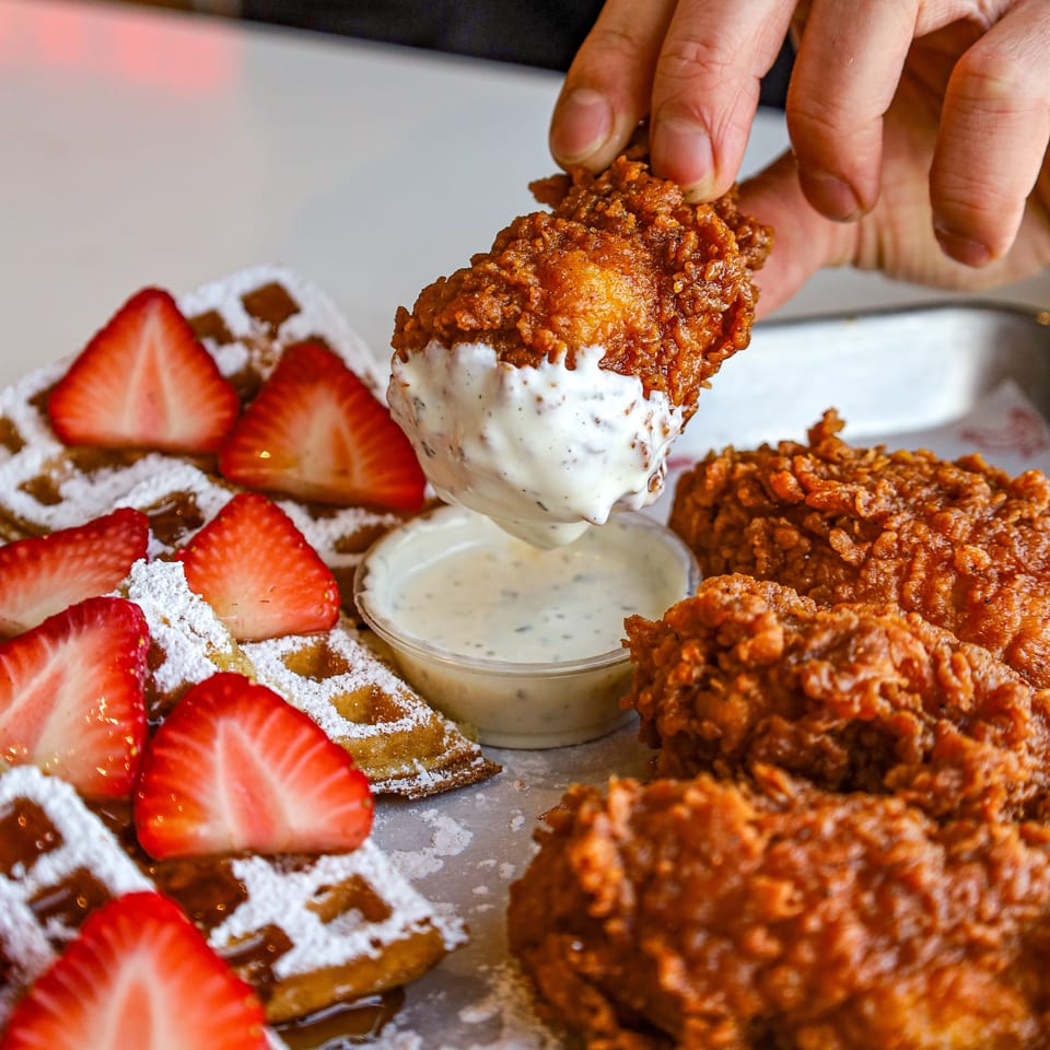 Hand dipping crispy fried chicken into sauce, served with waffles and fresh strawberries.