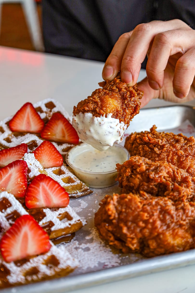 Hand dipping crispy fried chicken into sauce, served with waffles and fresh strawberries.