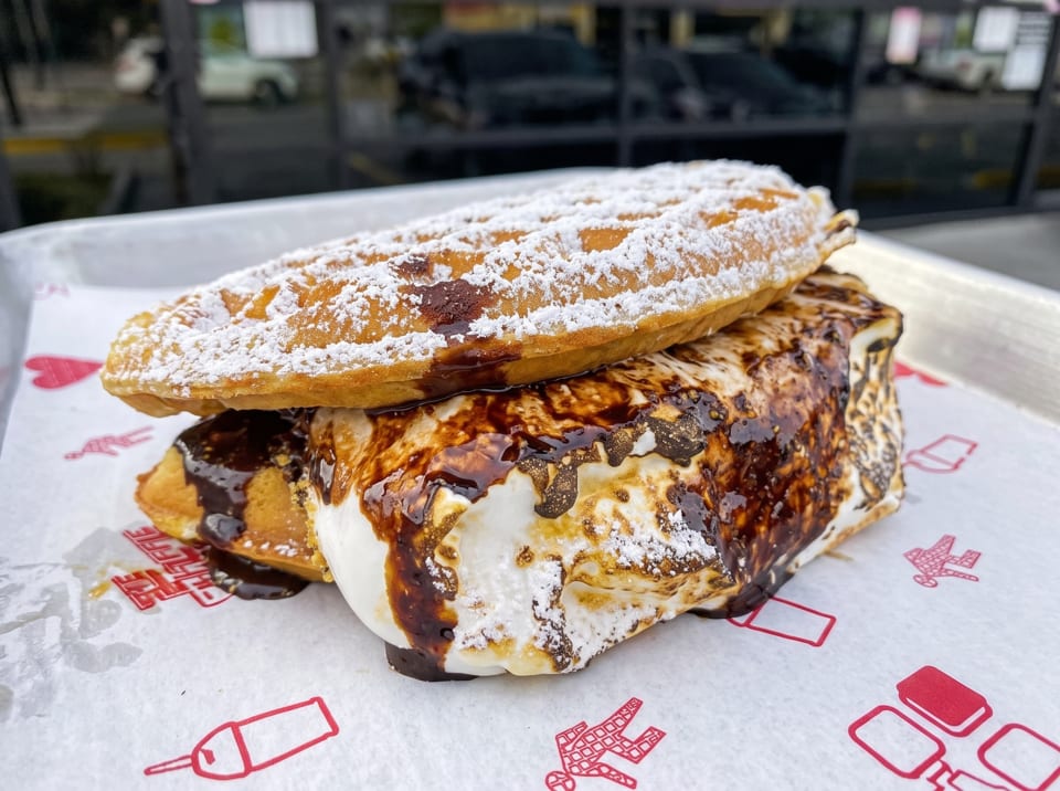 A waffle sandwich with toasted marshmallow, chocolate syrup, and powdered sugar on a branded paper.