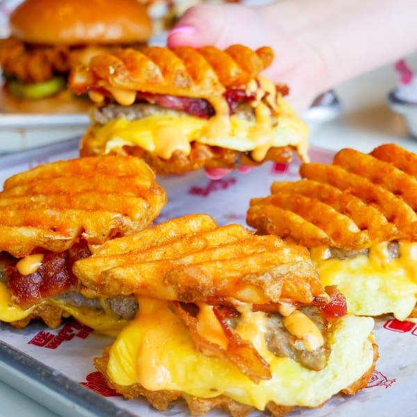 Several waffle-fry sandwiches with meat, cheese, bacon, and sauce on a branded tray.