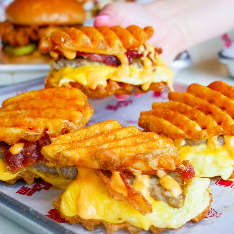 Several waffle-fry sandwiches with meat, cheese, bacon, and sauce on a branded tray.
