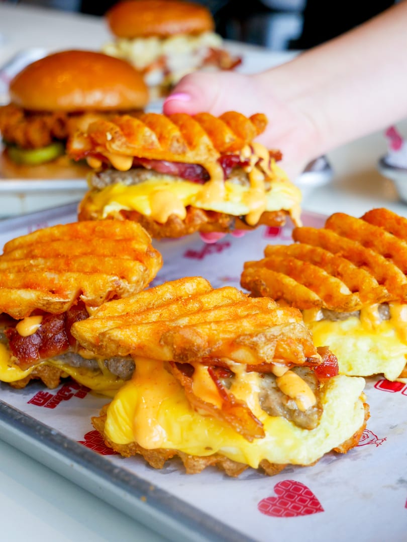 Several waffle-fry sandwiches with meat, cheese, bacon, and sauce on a branded tray.