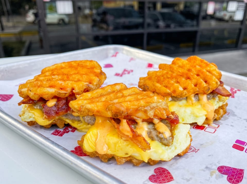 Three waffle fry sandwiches with egg, bacon, sausage, and sauce on a tray.