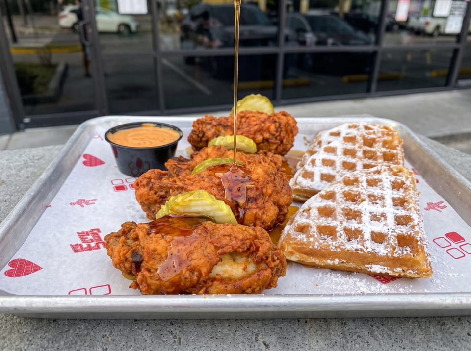 Honey being drizzled over fried chicken and waffles with pickles and sauce on a tray.