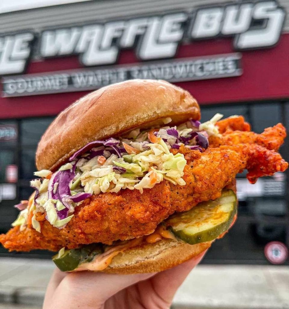 A hand holds a crispy chicken sandwich with coleslaw and pickles in front of The Waffle Bus.