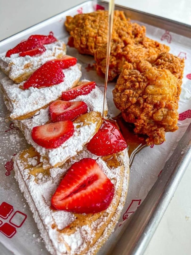 Golden fried chicken and powdered sugar waffles with strawberries, drizzled with syrup on a tray.